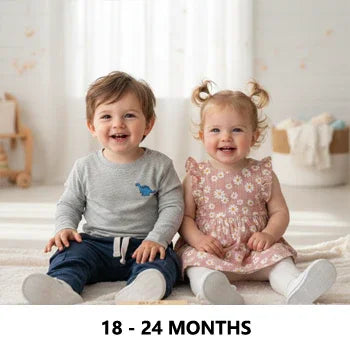 Two toddlers in Aatiyas Fashion apparel, one in a grey long-sleeve top and navy pants, the other in a pink floral dress, sitting indoors on a soft carpet, age 18-24 months.